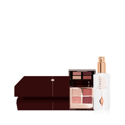 An open, dark brown mystery gift box with an open, quad eyeshadow palette with a mirrored lid with eyeshadows in shades of russet rose, dark brown, rose gold, and dull gold along with a face cream in a white-coloured bottle with a gold-coloured pump dispenser.