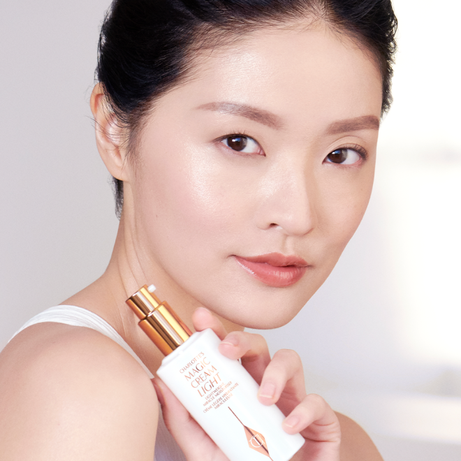 A fair-tone brunette model with luminous glass skin holding a face cream in a white-coloured bottle with a pump.