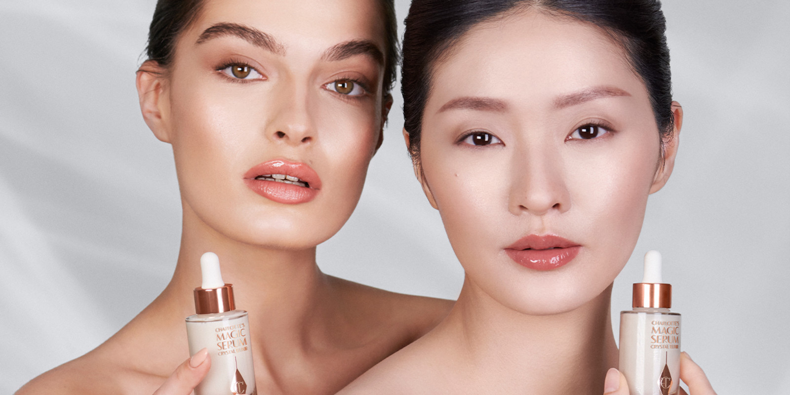 Two light-tone brunette models, one with mature skin, and both with dewy, glass skin, shoulders bare, and holding silver-coloured serum bottles.