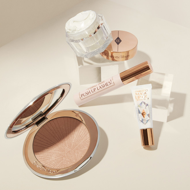 Open highlighter compact in a rose gold shade with a mirrored lid, pearly-white face cream in a glass jar with a gold-coloured lid, black-coloured mascara in a nude pink tube with a gold-coloured lid, and lip oil in a white-coloured tube with gold-coloured lid.