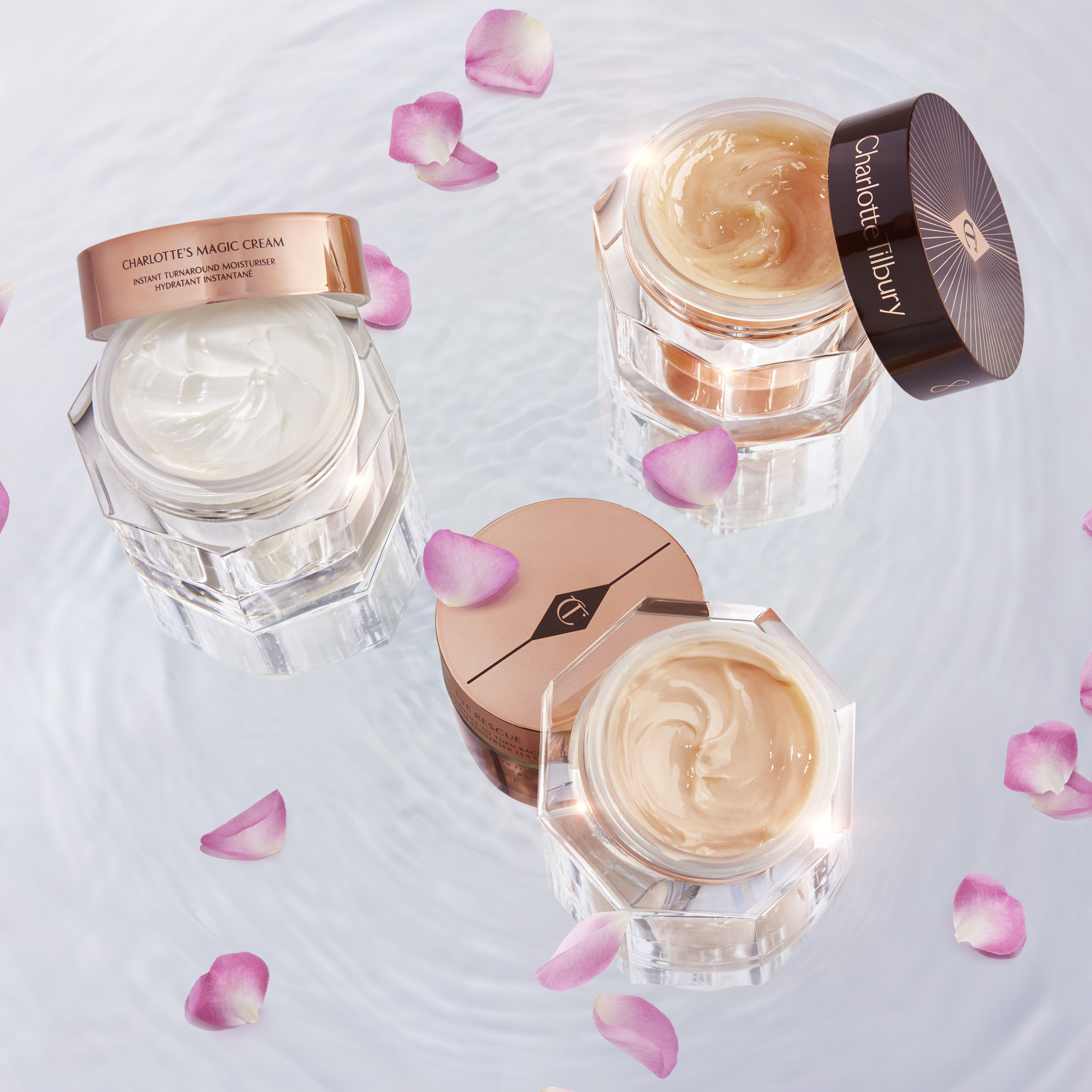 Pearly-white face cream in an open glass jar with a gold-coloured lid, peach-coloured thick night cream in an open glass jar with a dark-brown-coloured lid, and champagne-coloured eye cream in an open, petite glass jar with a gold-coloured lid, all three on top of crystal-clear water with rose petals falling down.
