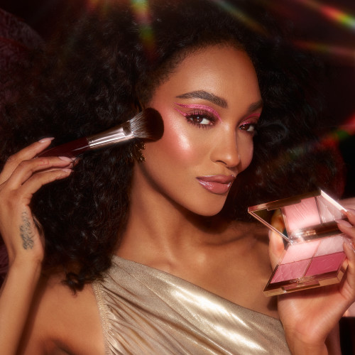 Deep-tone brunette model with brown eyes wearing full-glam, extremely glowy pink makeup while holding an open, face palette with matte and shimmery eyeshadows, blushes and highlighters in shades of pink and gold with a mirrored lid.