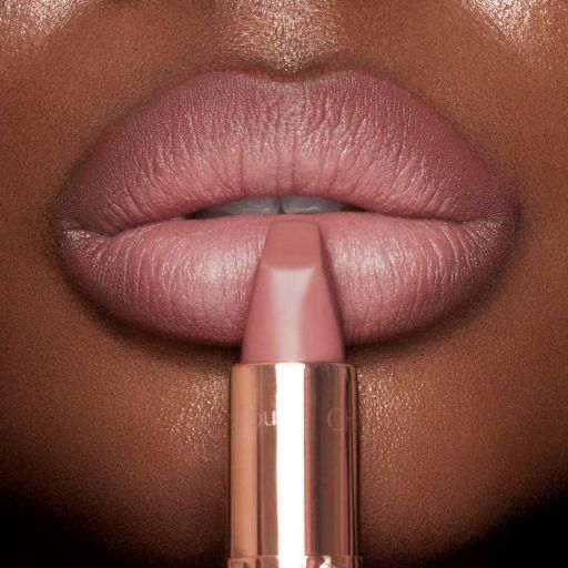A deep-skin model wearing a nude-pink lipstick and holding the lipstick, gently pressed to their lower lip. 