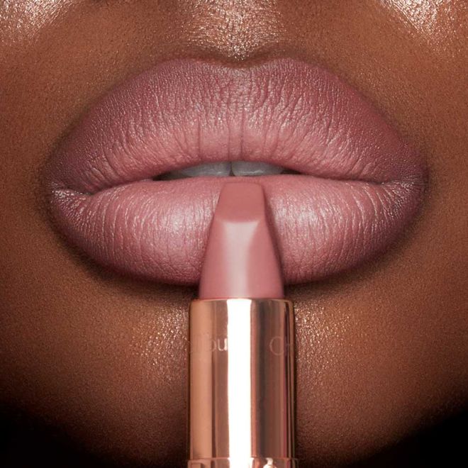Deep skin close-up of Matte Revolution lipstick in Pillow Talk Original A deep-skin model wearing a nude-pink lipstick and holding the lipstick, gently pressed to their lower lip.