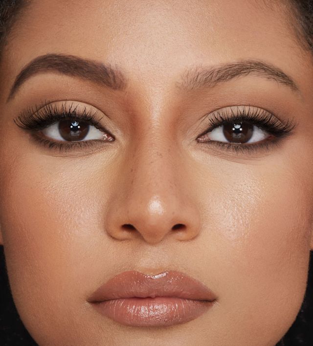 Close-up of a medium-tone model with brown eyes with one eyebrow completely bare and the other filled and fluffed, looking lush and full while also wearing black kohl pencil and nude lip gloss.