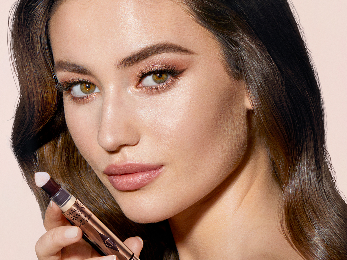 Light-toned model brunette model wearing dewy nude pink make-up with shimmery brown eye make-up and holding an open concealer.