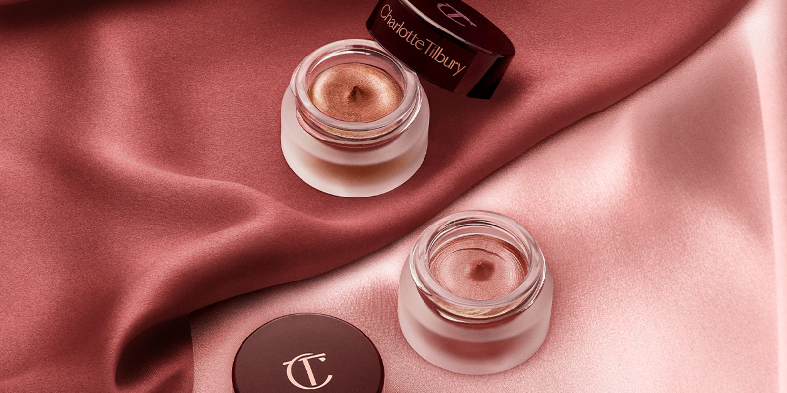 Two cream eyeshadows in glass pots, one a russet rose colour with a golden-peach sparkle and the other a nude pink with fine shimmer, with their lids placed next to them.