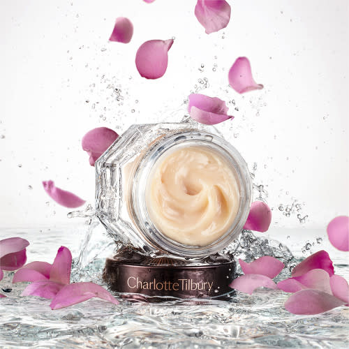 Banner with an open glass jar with thick, peach-coloured night cream and its dark-brown and gold-coloured lid placed on top of crystal-clear water with rose petals falling down on it.