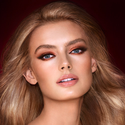 A light-tone model with blue eyes wearing smokey brown and gold eye makeup with warm pink blush and glossy nude-pink lips