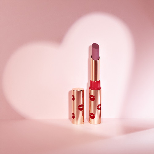 An open lipstick in a neutral rosy-pink colour with a matte finish with a gold-coloured tube and lid with red-coloured kiss print all over. 