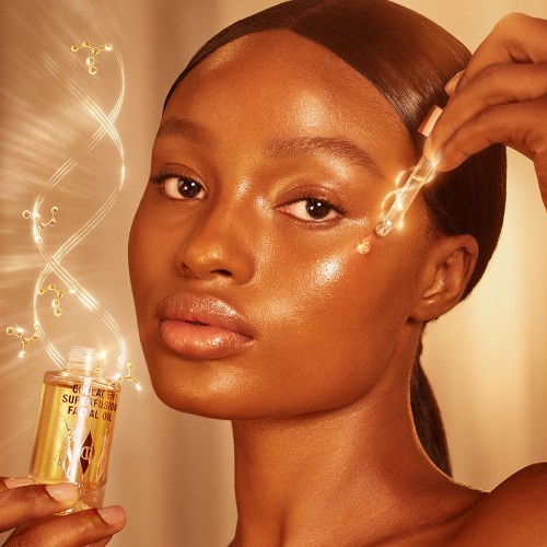 deep-tone model with glowy, luminous, glass skin, applying a light-gold-coloured facial oil from a glass bottle with a gold and white-coloured dropper lid.