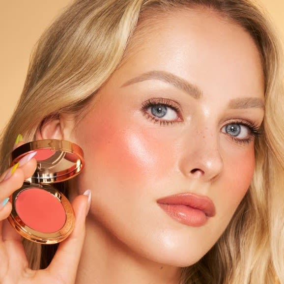 Blonde fair model wearing a peachy blush makeup look holding a peach cream blush.