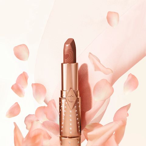 Banner with a lipstick with its lid removed in a nude peach shade in a gold-coloured tube with peach-coloured petals falling on it.