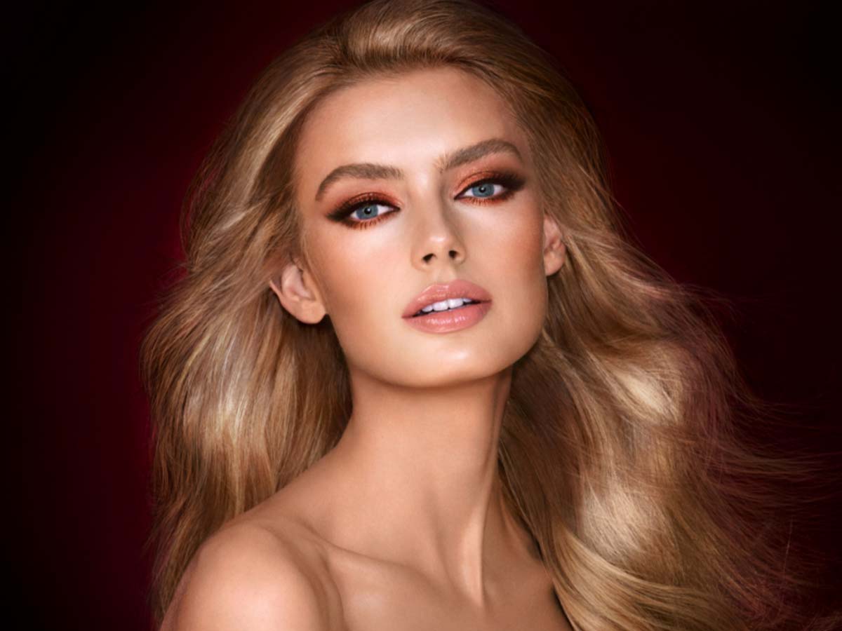 A light-tone model with blue eyes wearing smokey brown and gold eye makeup with warm pink blush and glossy nude-pink lips.