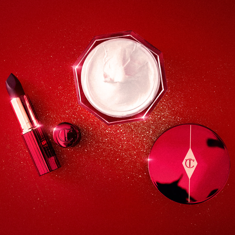A thick, pearly-white face cream in an open glass jar with an eye-catching red-coloured lid and an open lipstick in a dark purple shade with vivid red-coloured packaging. 