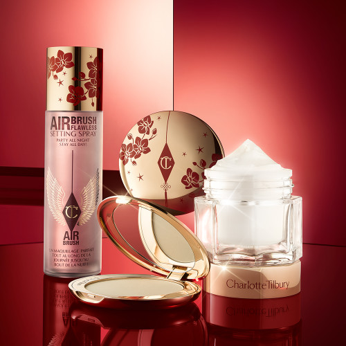 A matte burnt-orange red lipstick in gold-coloured packaging with cherry blossoms illustrated on the tube for the Lunar New Year, along with its lid next to it and an open, pressed powder compact for fair skin tones with a mirrored lid, in gold-coloured packaging with red-coloured cherry blossoms on the lid, setting spray with cherry blossoms on the lid, and a face cream in a glass jar with cherry blossoms on the lid.