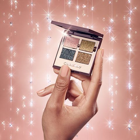 Close-up of a medium-tone model's hands holding an open, mirrored-lid quad eyeshadow palette with shimmery eyeshadows in rose gold, teal, honey-gold, and bright gold colours. 