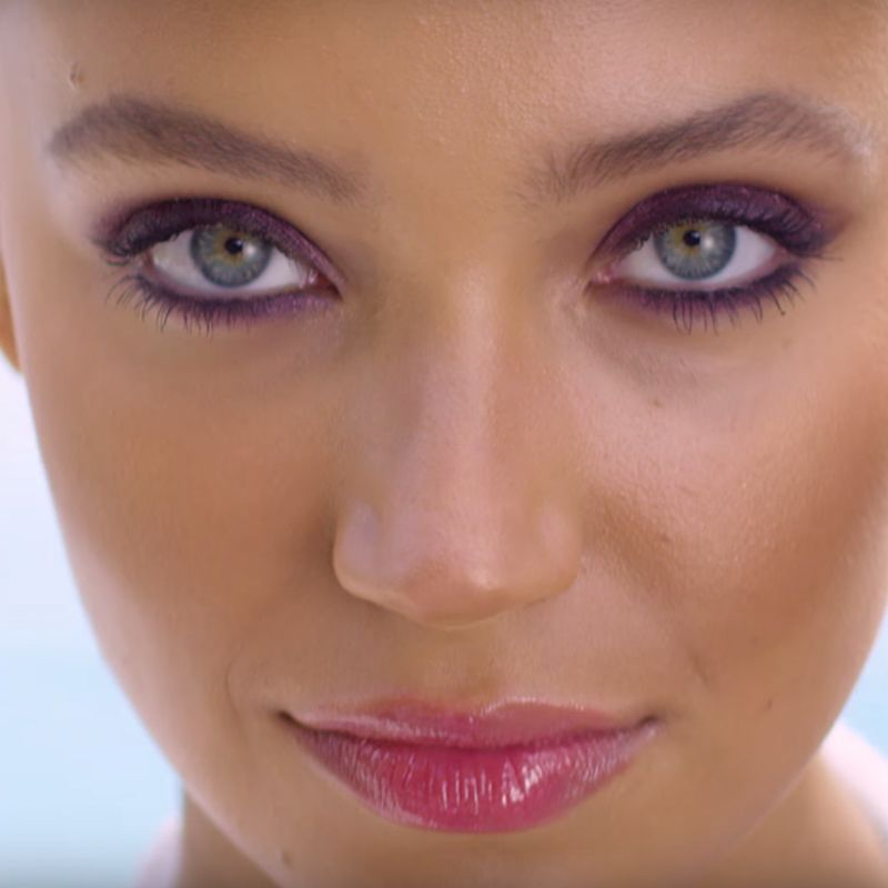 Close-up of a model with green eyes wearing shimmery, smokey purple eye makeup with a berry-pink lip gloss. 