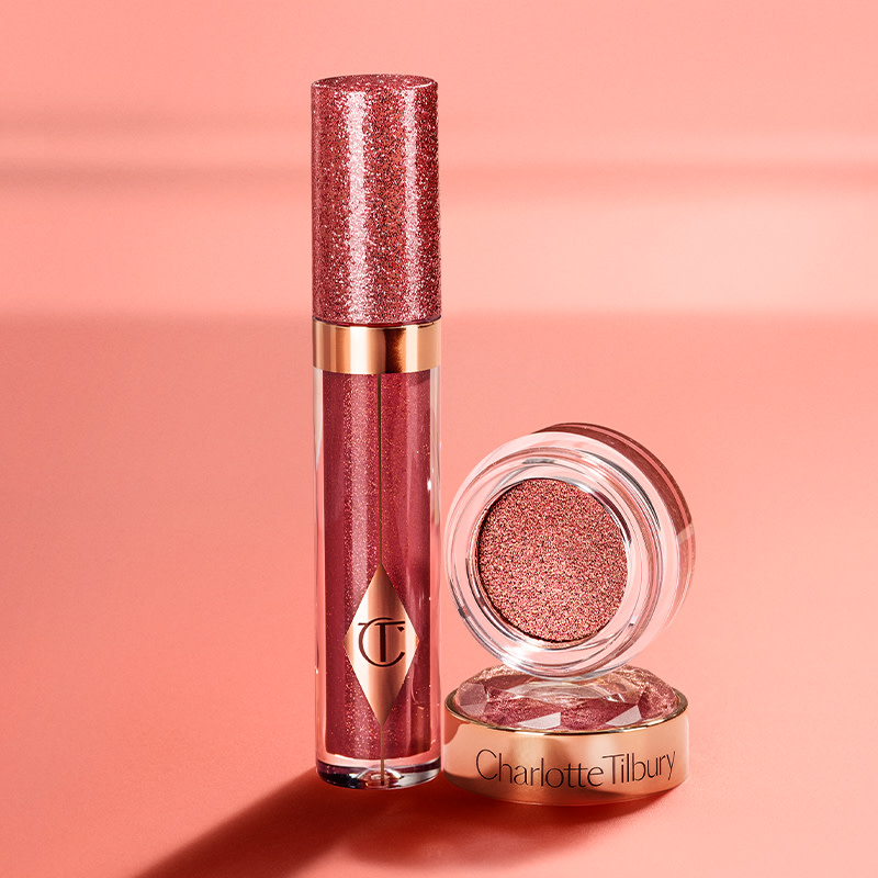 A peach-toned banner with a shimmery berry-pink lip gloss in a glass tube with a glittery lid next to shimmery, cream eyeshadow in a berry-pink shade in a glass pot with its lid removed.