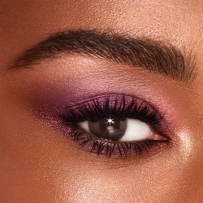 Single-eye close-up of a deep-tone model with brown eyes wearing shimmery purple, pink, and cream eye makeup. 