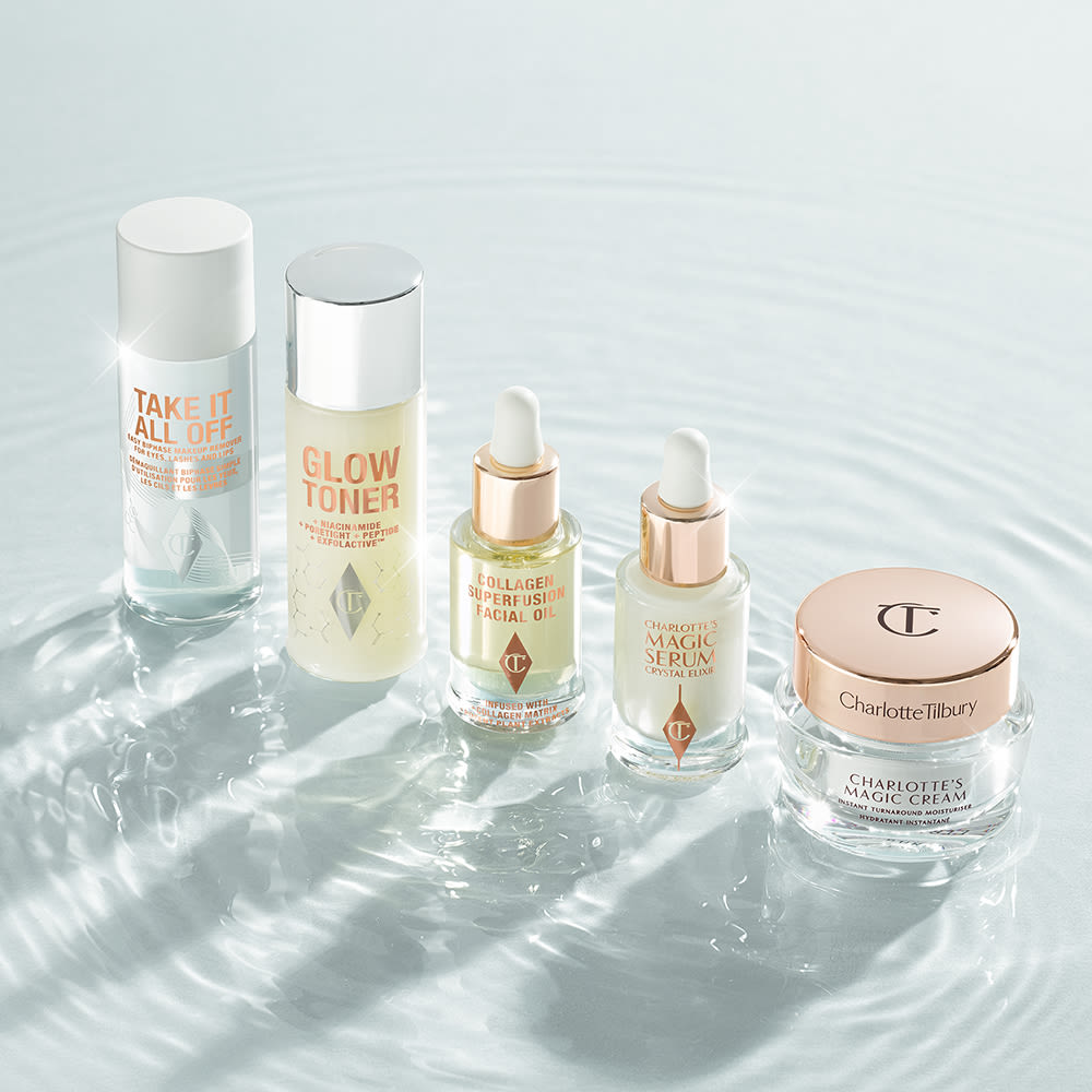 Travel-size, pearly-white face cream in a glass jar with a gold-coloured lid, makeup remover in a clear bottle with a white-coloured lid, toner in a clear bottle with a silver-coloured lid, light gold-coloured face oil in a glass bottle with a dropper lid, and luminous face serum in a glass bottle with a white and gold dropper lid.