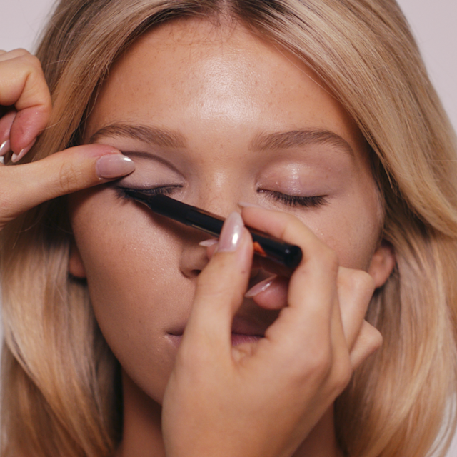 Makeup artist applying black liquid eyeliner close to the upper lash line of a fair blonde model.