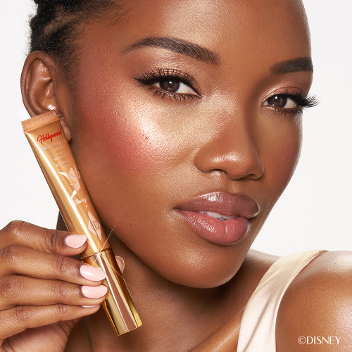 Deep skinned model wearing a glowy makeup look and holding Charlotte Tilbury's Disney liquid highlighter in Gold.