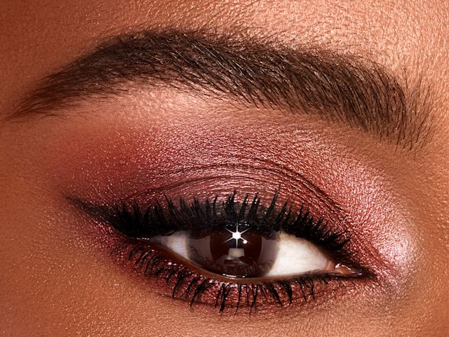 Single-eye close-up of a deep-tone model with brown eyes wearing shimmery red and rose-gold eyeshadow. 