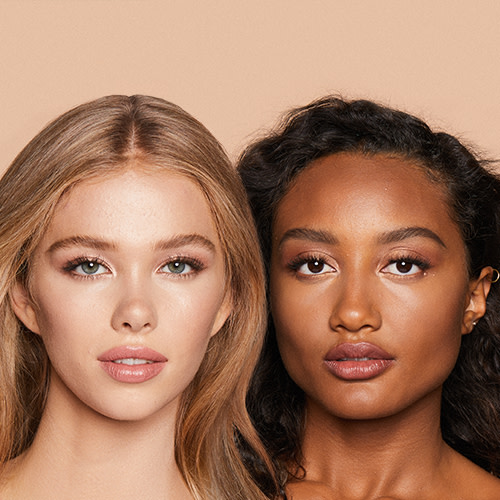 A fair-tone blonde model and a deep-tone brunette model with glowy, flawless face base, wearing golden-peach lipstick with bright champagne and fawn eye makeup. 