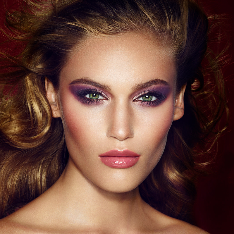 A light-tone model with blue eyes wearing smokey plum eye makeup with warm pink blush and glossy berry-pink lips 