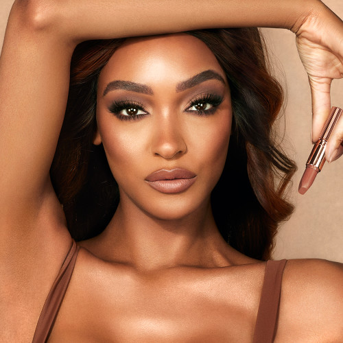 Deep-tone model with brown eyes wearing soft beige and champagne eyeshadow with a fresh, neutral nude peach matte lipstick while holding that lipstick up.