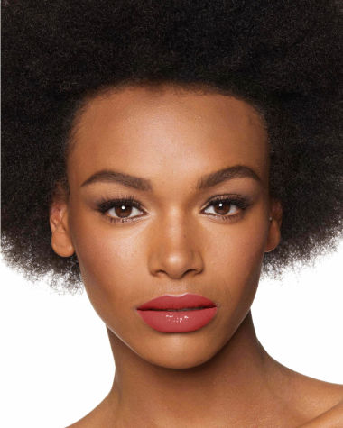 Deep-tone model with brown eyes wearing a moisturising lipstick balm in a soft glossy coral shade with a high-shine finish.
