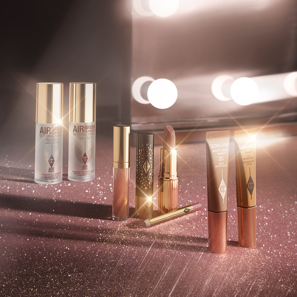 Collection of mini makeup products, which includes setting sprays in clear bottles with gold-coloured lids, lip glosses, lipsticks, highlighter wands, blush wands, and lip liner pencils.