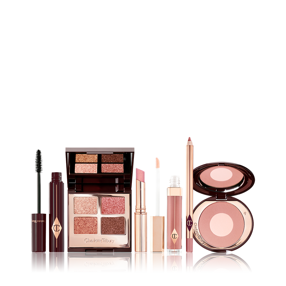 An open mascara tube with the applicator next to it with an open, mirrored-lid quad eyeshadow palette in shimmery neutral shades, a shimmery nude pink lipstick, a nude pink lip gloss and lip liner pencil, and a two-tone powder blush compact in a nude pink shade. 