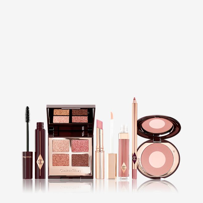 Pillow Talk Fresh, Glowing Makeup Secrets Box open An open mascara tube with the applicator next to it with an open, mirrored-lid quad eyeshadow palette in shimmery neutral shades, a shimmery nude pink lipstick, a nude pink lip gloss and lip liner pencil, and a two-tone powder blush compact in a nude pink shade.