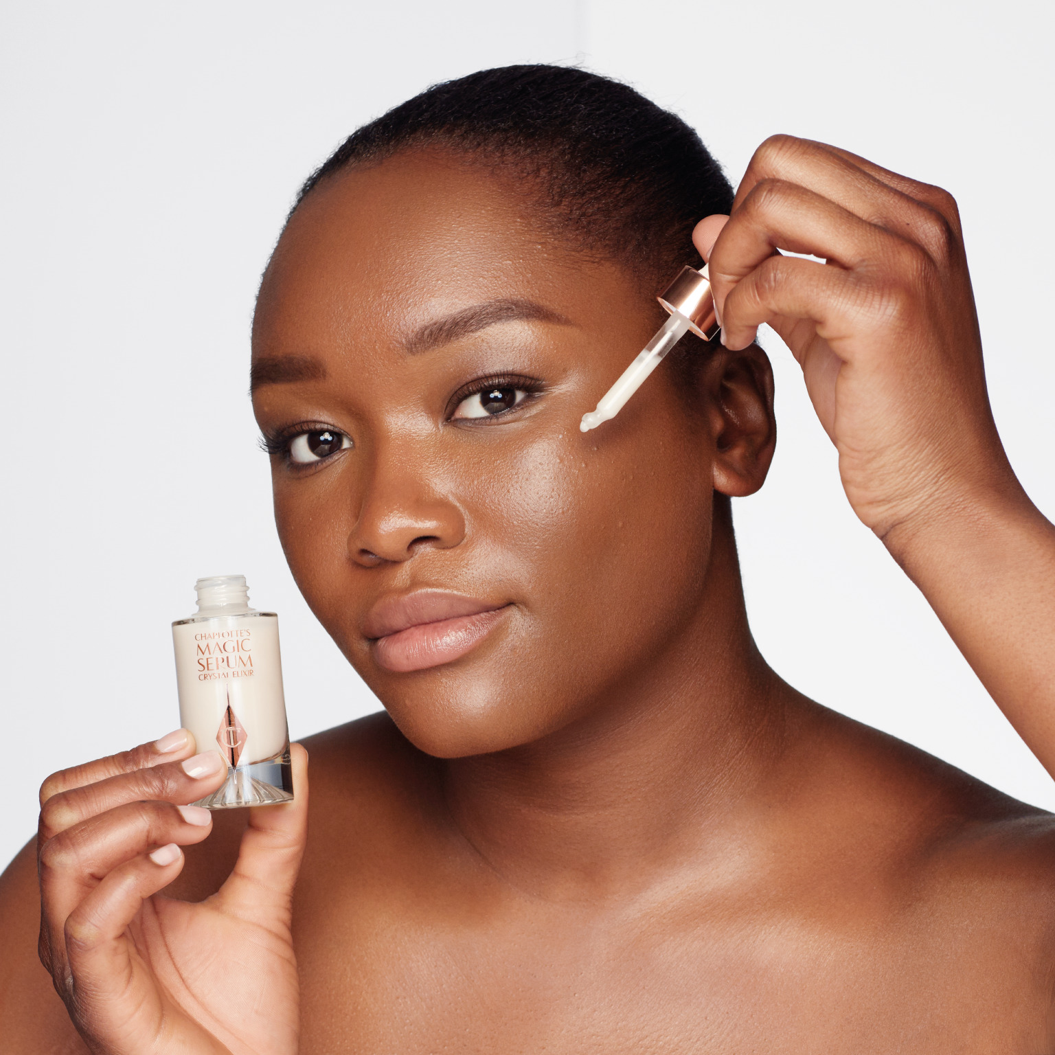 A deep-tone model with brown eyes wearing subtle eye makeup and applying a luminous face serum on her face with a gold-coloured dropper. 