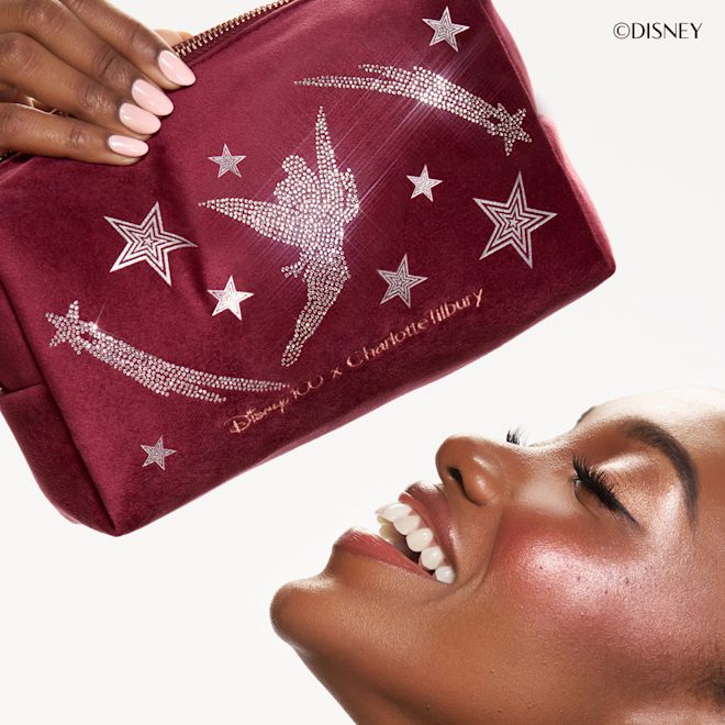 Model looking at Charlotte Tilbury Disney makeup bag Close up of deep skinned model looking up and laughing, holding the Charlotte Tilbury Disney100 velvet Tinker Bell makeup bag.