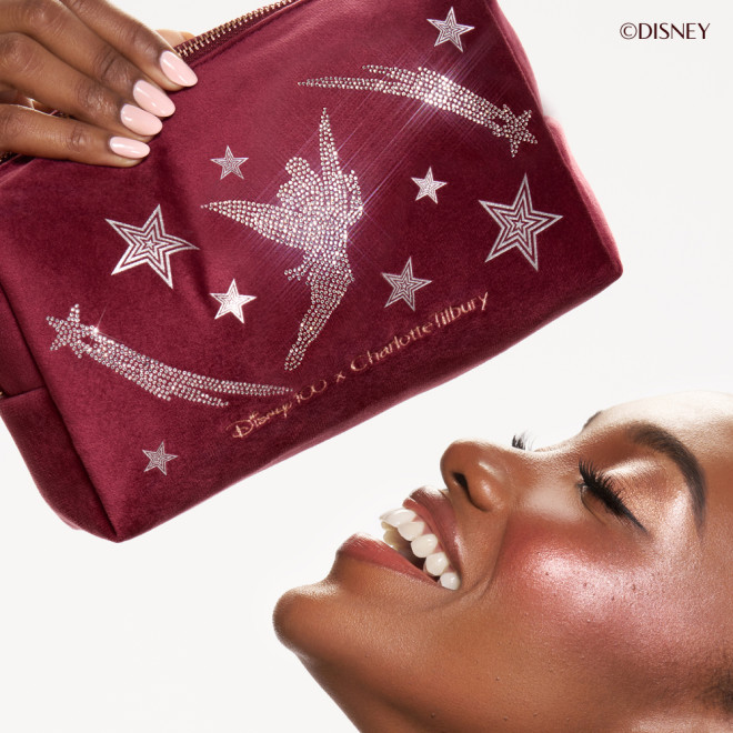 Close up of deep skinned model looking up and laughing, holding the Charlotte Tilbury Disney100 velvet Tinker Bell makeup bag.