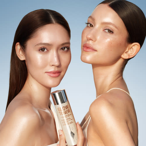 Two, fair-tone models with glowing, fresh skins holding a large toner bottle filled with a creamy, silvery-white-coloured toner with a silver-coloured lid. 