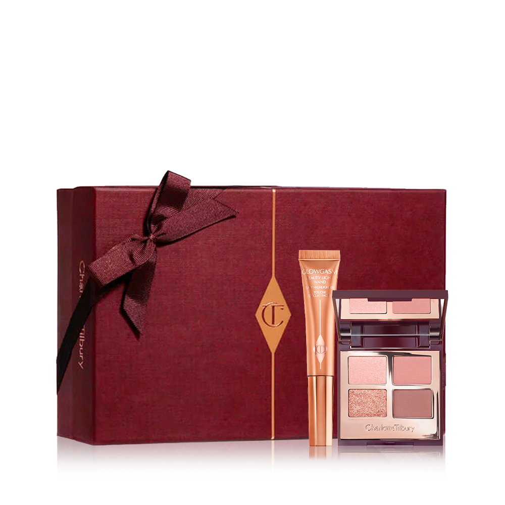 Liquid highlighter wand in a peach-gold-coloured tube with an open, mirrored-lid eyeshadow palette with matte and shimmery eyeshadows in brown, pink, and gold shades with a dark crimson-coloured gift box in the back. 