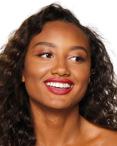 A deep-tone brunette model with brown eyes wearing shimmery beige and cream-coloured eyeshadow with a vibrant, poppy-red lip tint. 