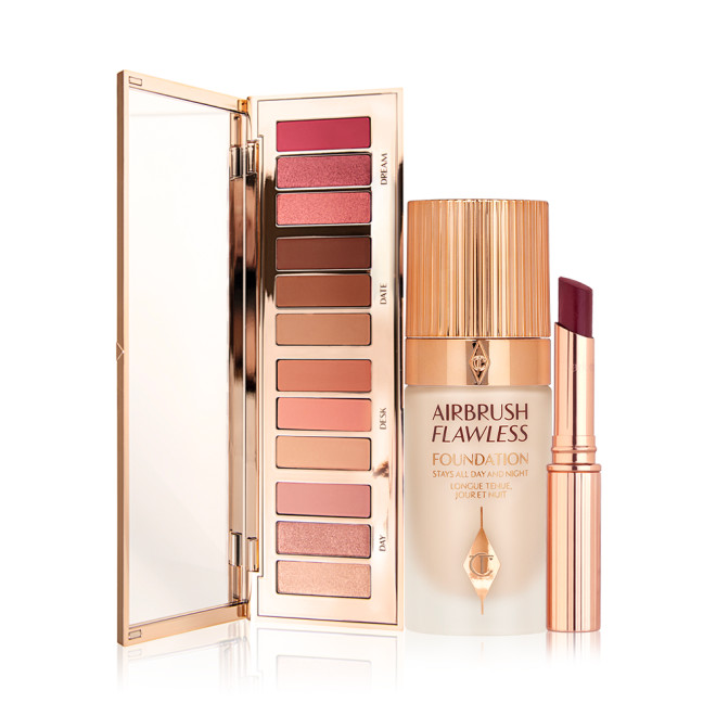 An open, mirrored-lid eyeshadow palette with matte and shimmery shades of pink, brown, peach, and gold, foundation in a frosted glass bottle with a gold-coloured lid, and a berry glossy lipstick in a gold-coloured tube. 