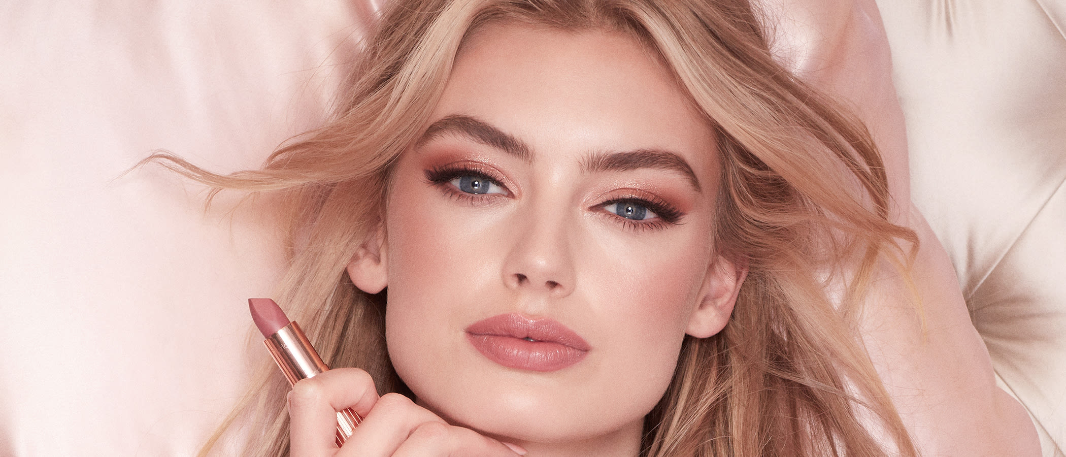 A fair-tone model with blonde hair and blue eyes wearing youthful, dewy, nude pink makeup while holding a nude-pink matte lipstick. 
