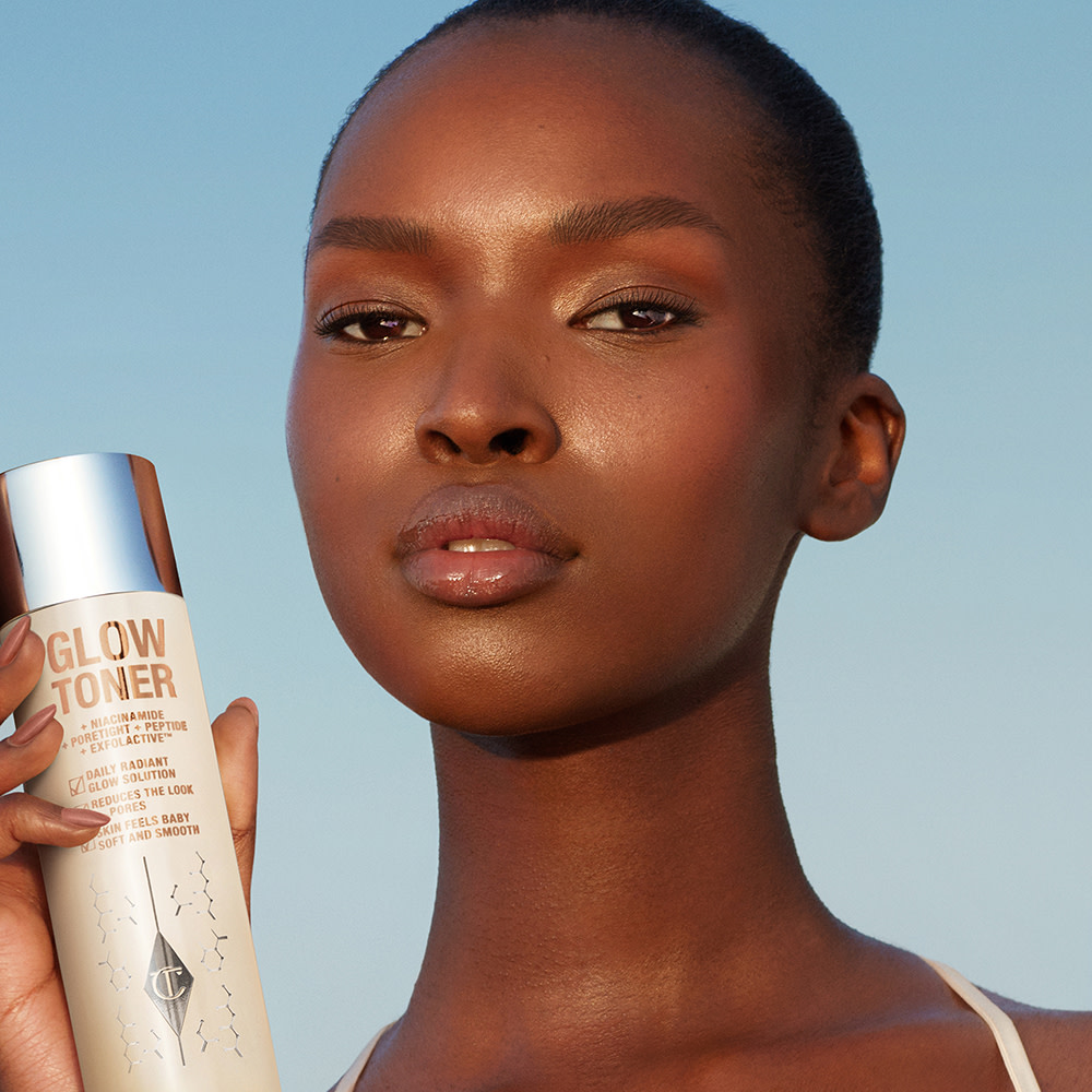 Deep-tone model with glowing, fresh skin holding a large toner bottle filled with a creamy, silvery-white-coloured toner with a silver-coloured lid. 