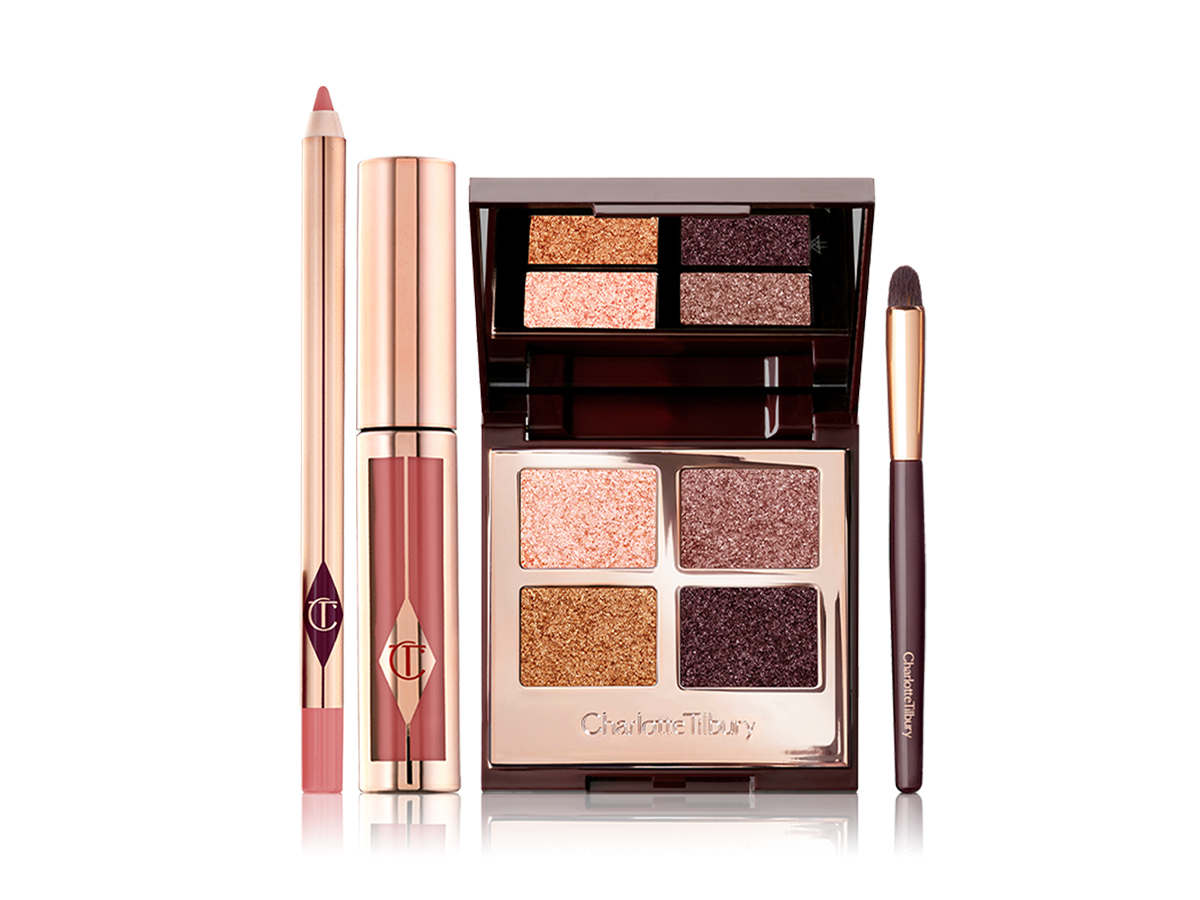 A nude pink lip liner pencil, liquid lipstick in a nude pink shade in gold coloured packaging, an eyeshadow smudger brush, and an open, quad eyeshadow palette with a mirrored-lid with four shimmery eyeshadows in shades of gold, pink, and grey.