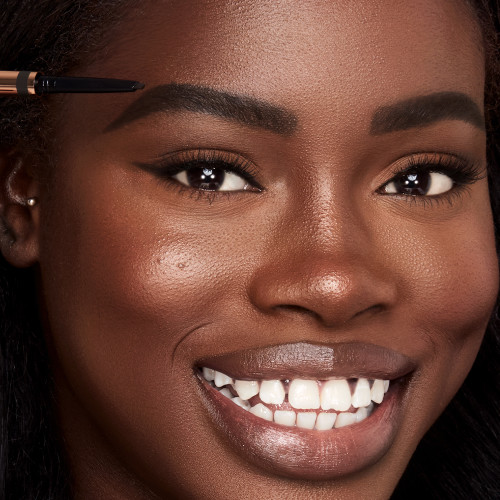 Deep-tone model with brown eyes filling in her brows with a black-coloured eyebrow pencil.