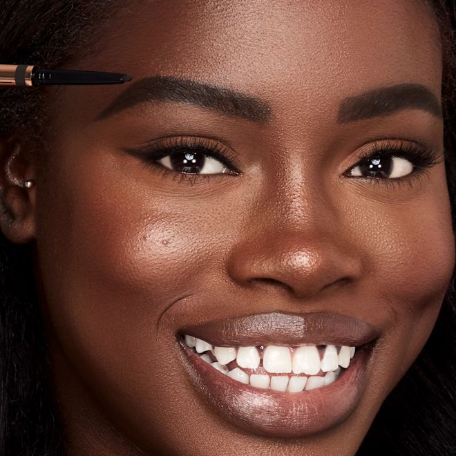 Brow Cheat in Natural Black on deep-tone model close-up Deep-tone model with brown eyes filling in her brows with a black-coloured eyebrow pencil.