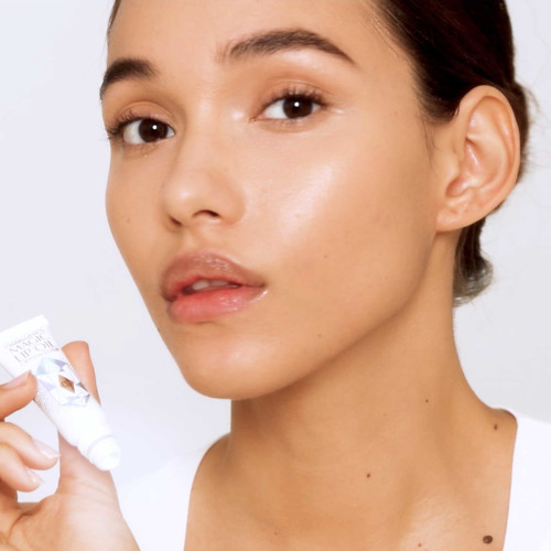 A light-tone brunette model with flawless glass skin and smooth, plump lips holding a tube of lip oil in white-coloured packaging. 
