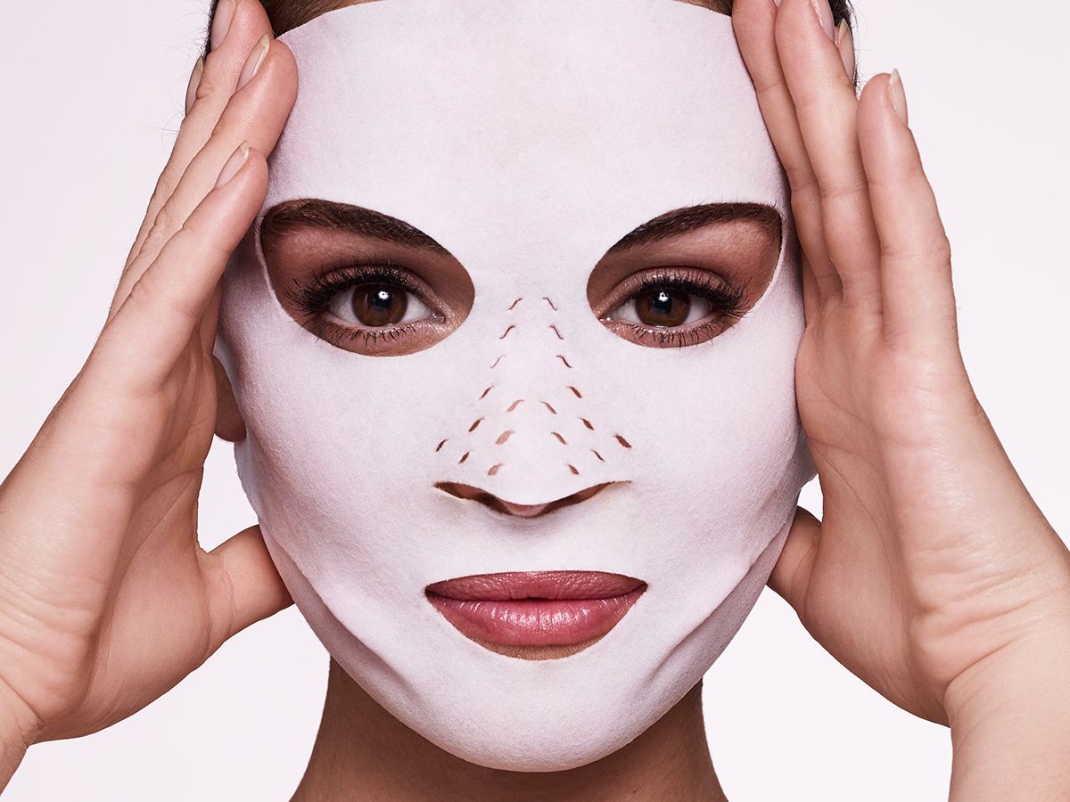 Close-up of a light-tone, brunette model wearing a sheet mask made of textile fabric with eyes, lips, and nose openings. 