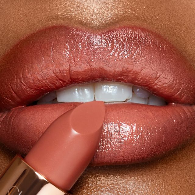 Lips close-up of a deep-tone model wearing a warm coral lipstick with a satin-finish while holding the lipstick in front of her lips.
